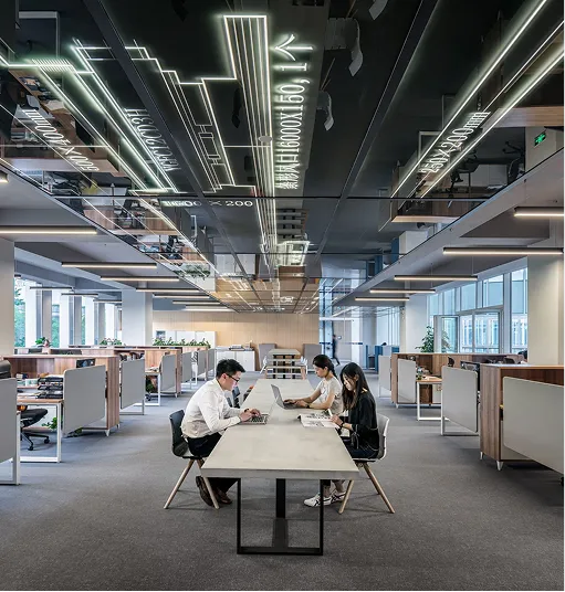 Modern office with three coworkers seated at a long table working on laptops and documents, surrounded by cubicles and large windows.