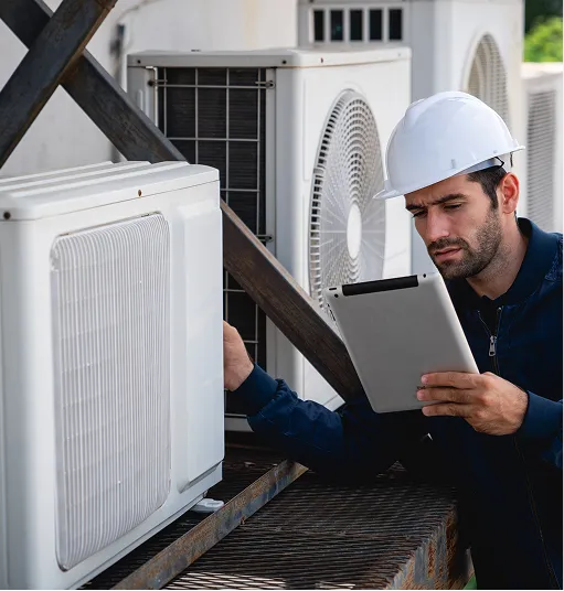 Technician in a white hard hat inspecting an outdoor HVAC unit while holding a tablet.