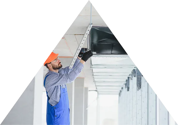 Worker in blue overalls and orange helmet installing or inspecting metal ductwork on a ceiling.