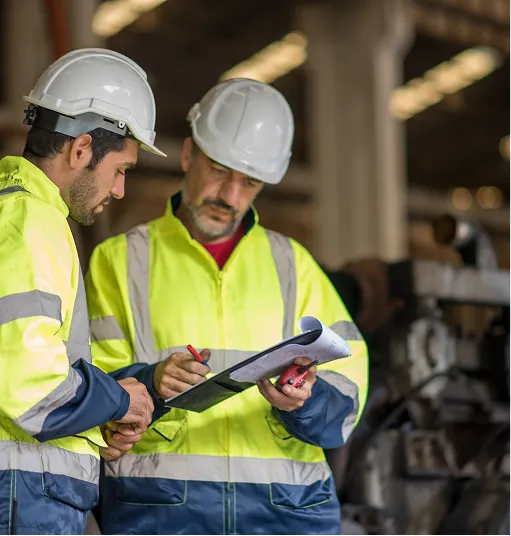 Two construction workers in white helmets and yellow safety jackets reviewing documents in an industrial setting.
