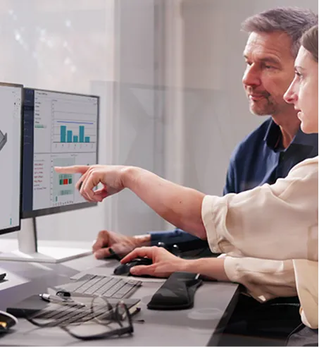Two professionals examining data charts on a dual computer monitor setup in an office.