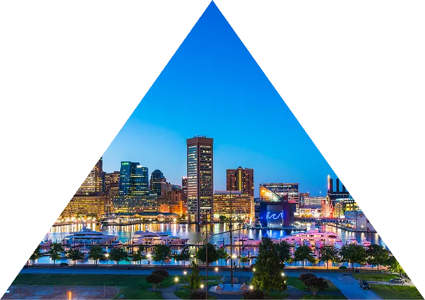 Evening skyline of Baltimore featuring illuminated buildings and a waterfront marina with boats under a clear blue sky.