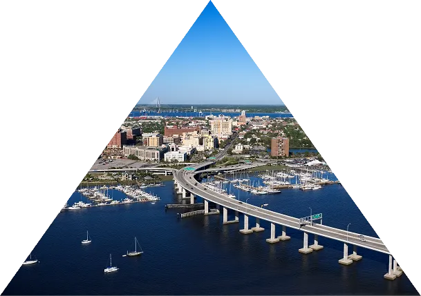 Aerial view of a curved bridge over water leading to a city with boats docked nearby under a clear blue sky.