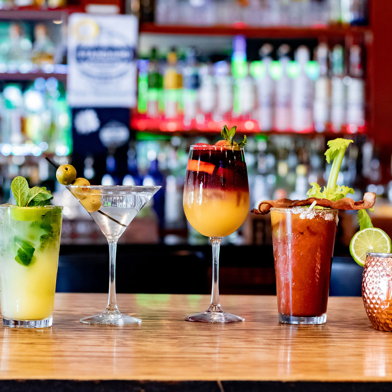 A bar counter displays five colorful cocktails, including a mojito, martini, mimosa, Bloody Mary, and a copper-mugged drink, set against a vibrant bar backdrop.
