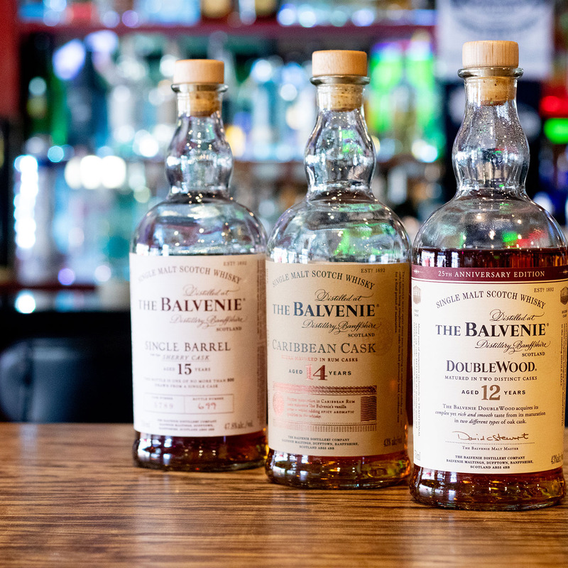 Three bottles of Balvenie Scotch whisky on a wooden bar. Labels read: Single Barrel 15, Caribbean Cask 14, Doublewood 12. Bar lights softly blur in the background.