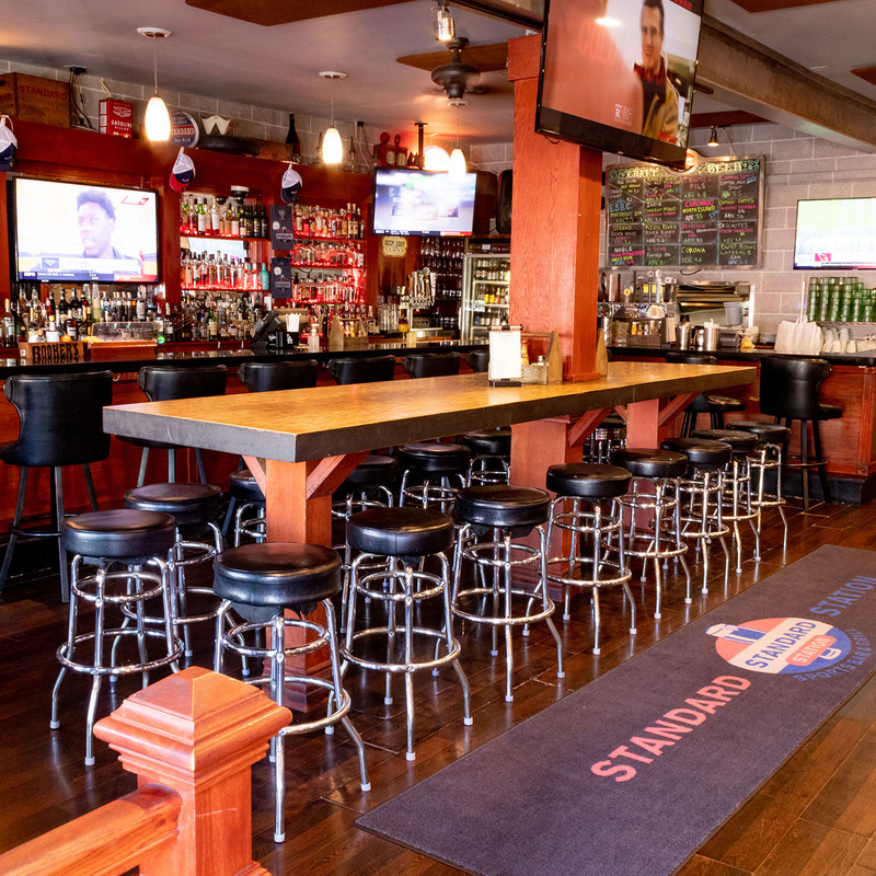 Cozy bar interior with wooden accents, featuring a long communal table and bar stools. Multiple TVs display sports.