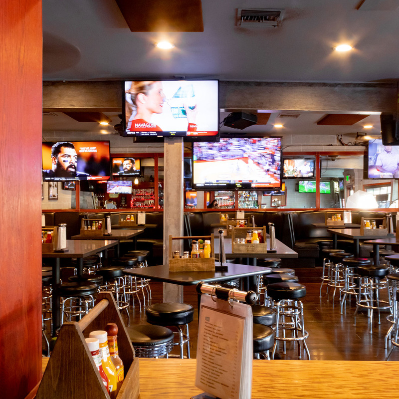 Lively sports bar interior with multiple screens displaying various channels. Bar stools and tables fill the room, condiments neatly arranged. Cozy and inviting ambiance.