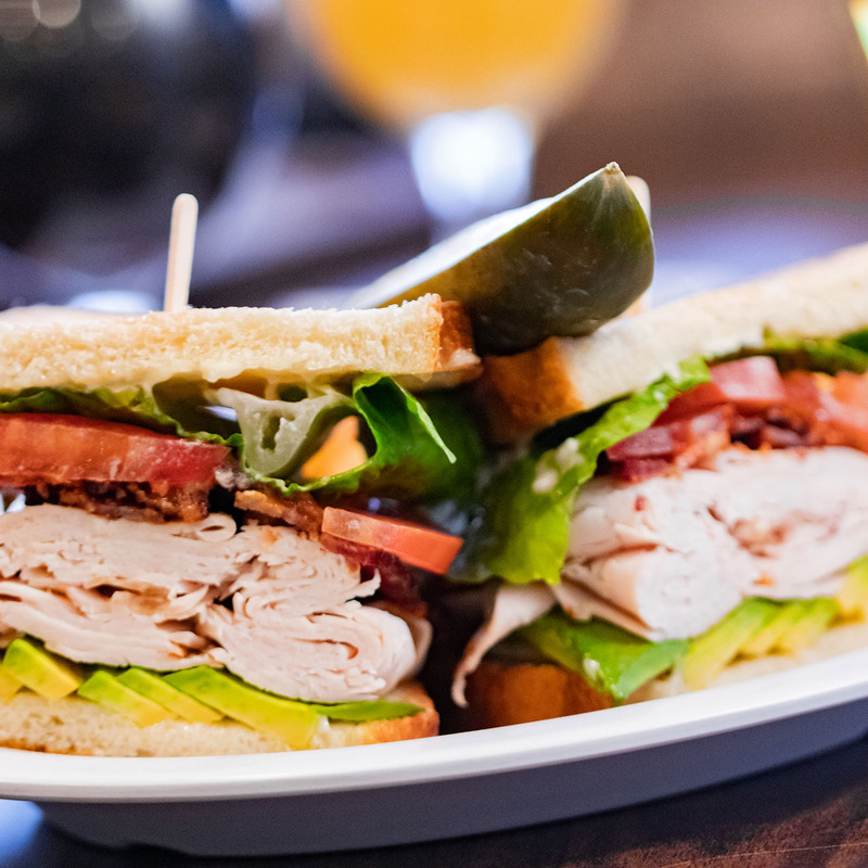 Close-up of a fresh turkey sandwich on white bread with lettuce, tomato, bacon, avocado, and a pickle on top. Served on a white plate.