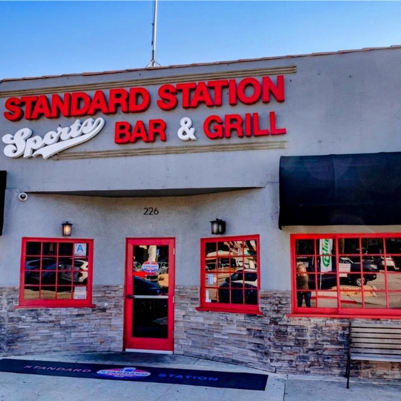 Facade of STANDARD STATION SPORTS BAR & GRILL with a vintage feel. Red-framed windows and door, stone wall accents, and bold red signage.