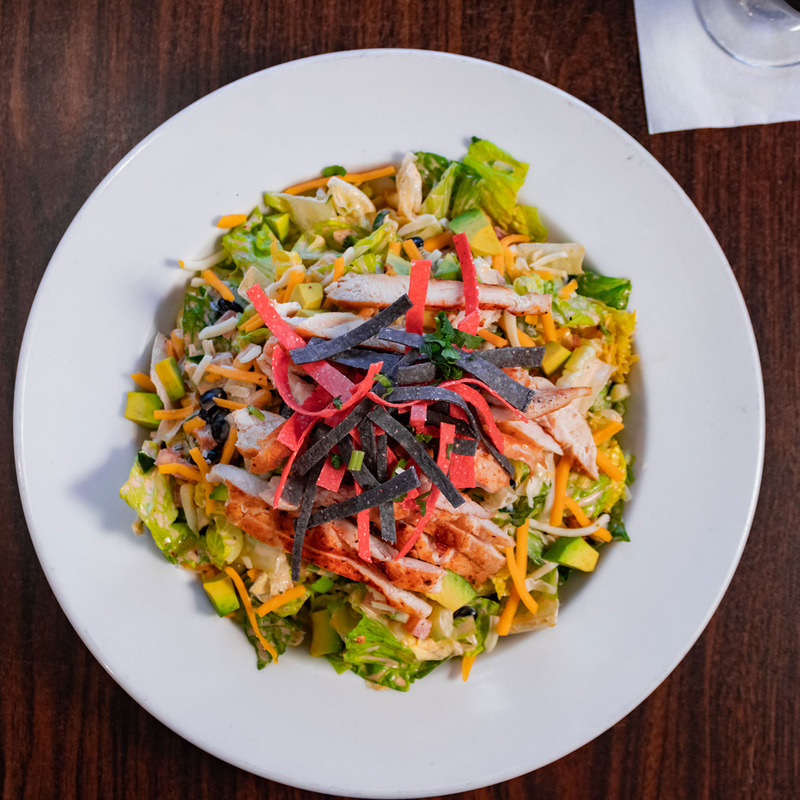 A colorful salad with lettuce, shredded cheese, avocado, grilled chicken, and red and black tortilla strips on a white plate on a wooden table.