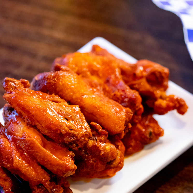 Close-up of crispy, golden-brown buffalo chicken wings on a white plate, placed on a wooden table. The wings are glazed with a vibrant orange sauce.