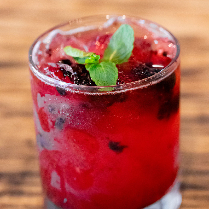 A refreshing red berry cocktail with ice cubes in a glass, garnished with fresh mint leaves on top, sitting on a wooden table background.