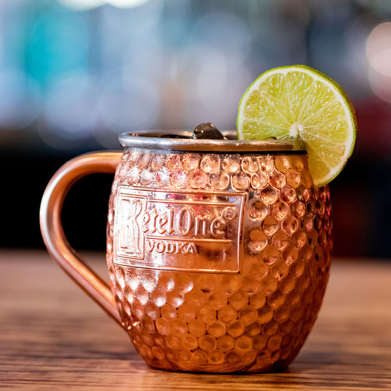 Copper mug on a wooden table, filled with ice and garnished with a lime slice. Features embossed logo, with bokeh lights in the blurred background.
