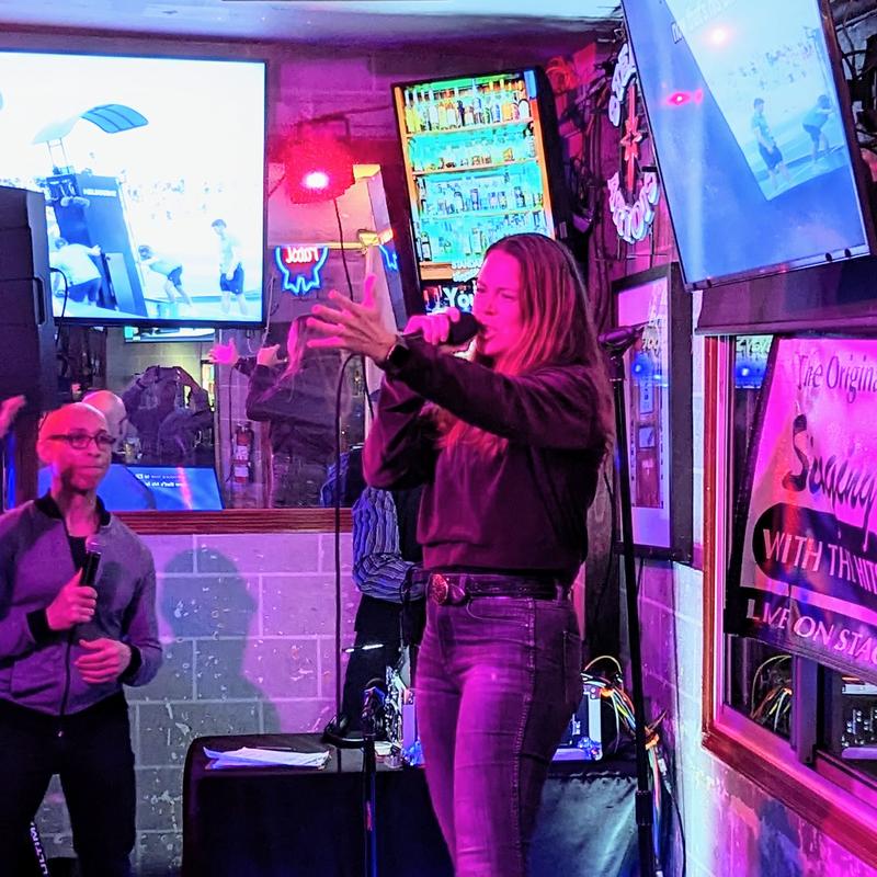 A woman sings into a microphone at a lively karaoke bar. Bright neon lights and TV screens create an energetic atmosphere.