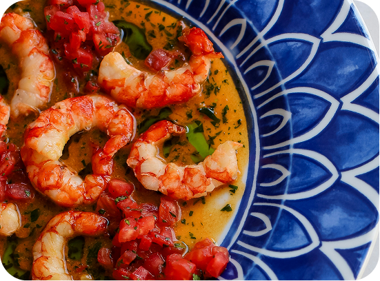Cooked shrimp in a seasoned sauce with diced tomatoes served in a blue and white patterned bowl.