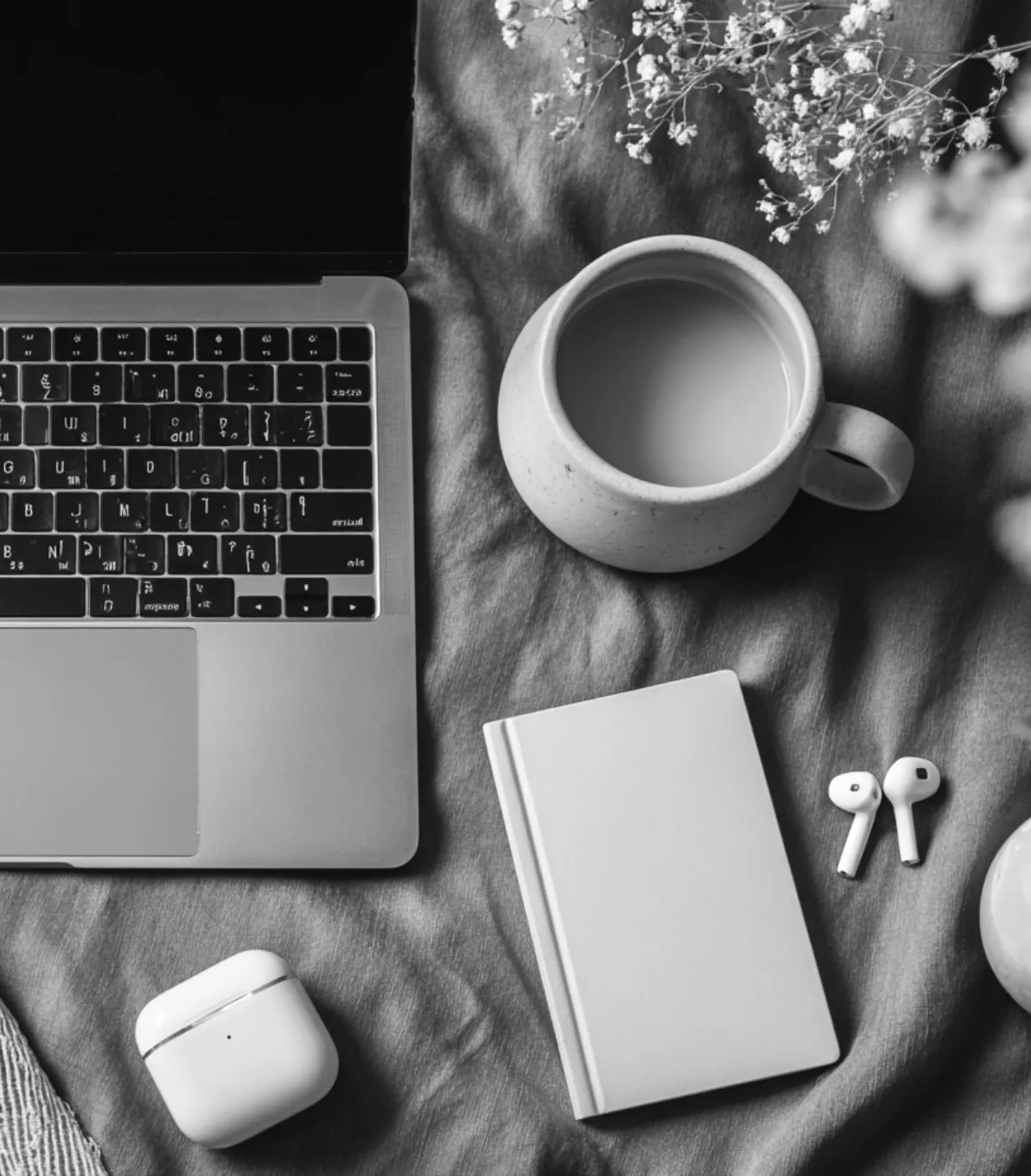 Flat lay of a laptop, coffee cup, closed book, wireless earbuds, and earbud case on a fabric surface with flowers.