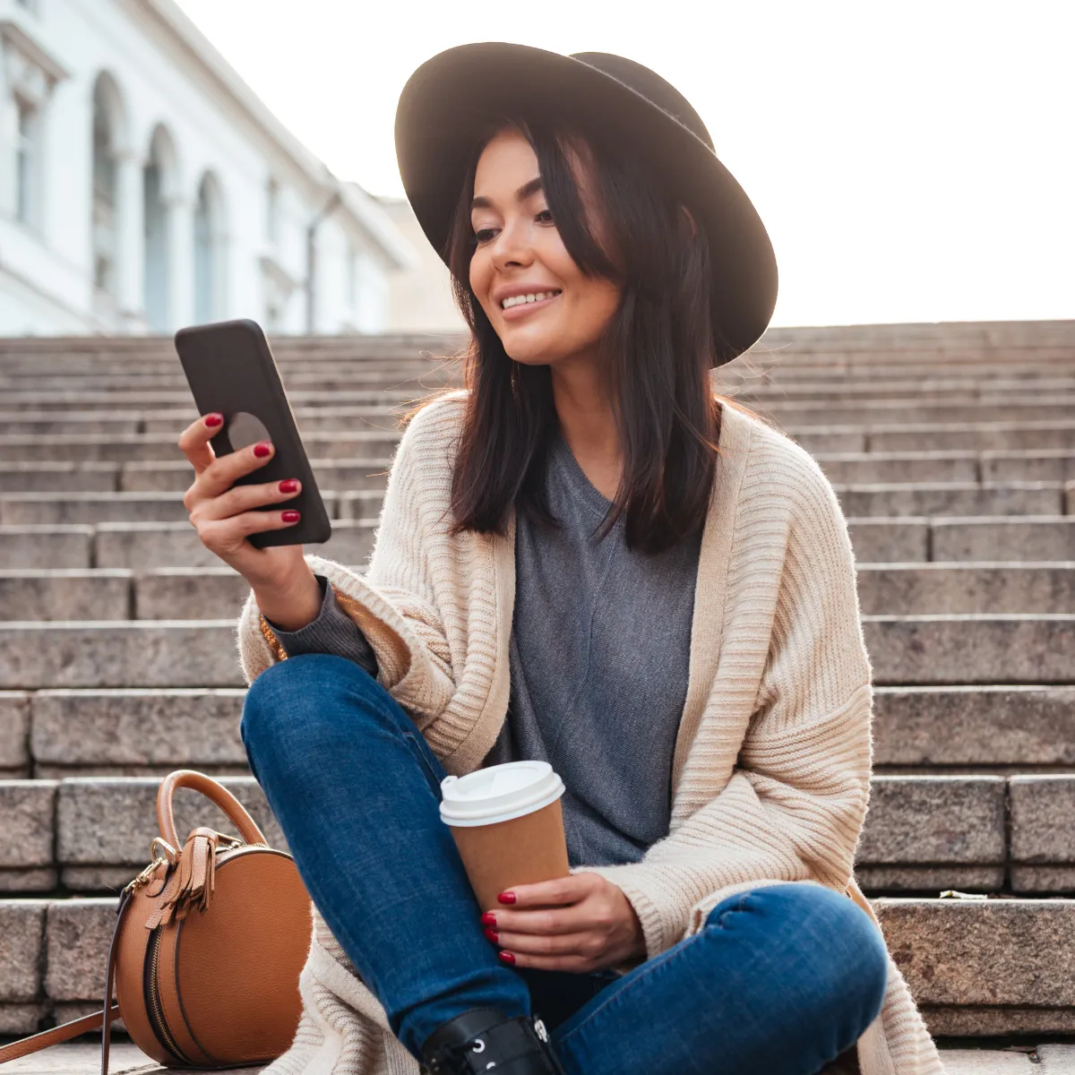 Smiling woman wearing a black hat and beige cardigan sitting on stone steps, holding a smartphone and a coffee cup with a tan bag beside her.