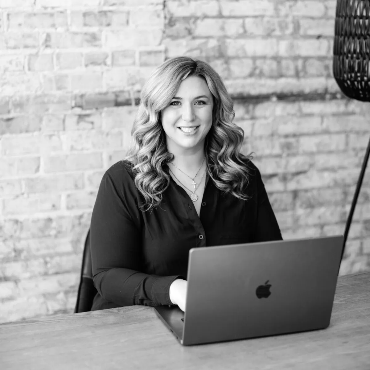 Smiling woman with wavy hair sitting at a desk working on a laptop in front of a brick wall.