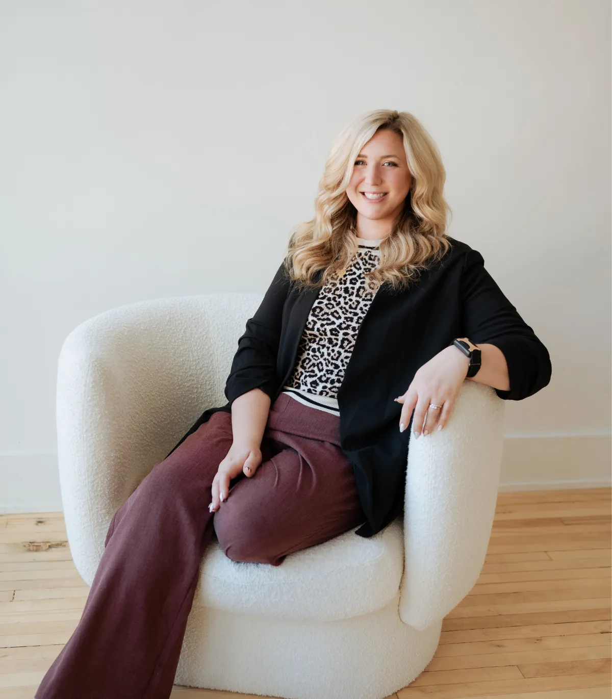 Smiling blonde woman with wavy hair wearing a leopard print top, black blazer, and maroon pants sitting on a white armchair.