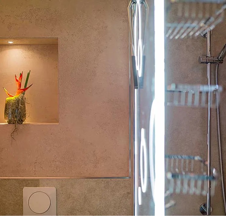 Beleuchtete Wandnische im Badezimmer mit einer Pflanze und Vogel-des-Paradieses-Blüten neben einem Duschbereich mit Duschkorb und verchromtem Duschkopf.