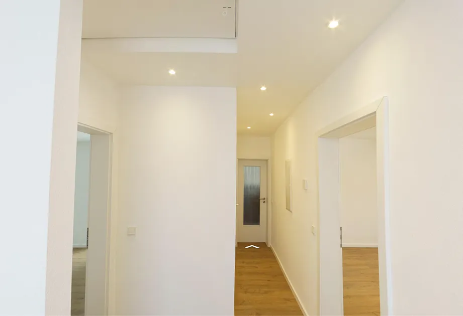 Heller Flur mit weiß gestrichenen Wänden, Holzboden und mehreren offenen Türen sowie einer geschlossenen Tür am Ende.