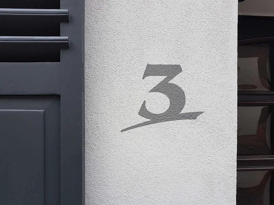 Graue Hausnummer 3 auf einer weißen Putzwand neben einer schwarzen Tür.