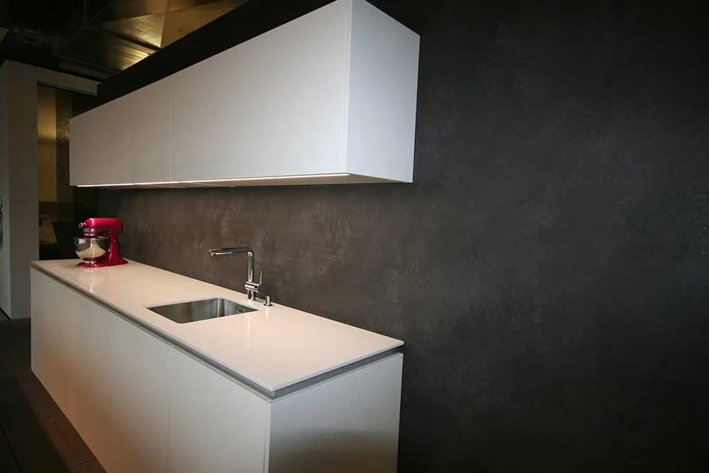 Moderne weiße Küche mit weißer Arbeitsplatte, integriertem Spülbecken, rotem Küchenmixer und dunkler Wand im Hintergrund.