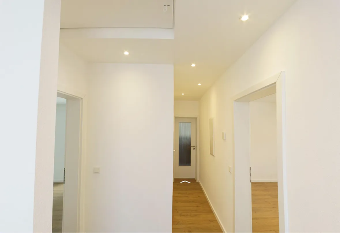 Flur mit weißen Wänden, Holzboden und eingebauten Deckenleuchten, zwei Türöffnungen auf beiden Seiten und eine Tür am Ende.
