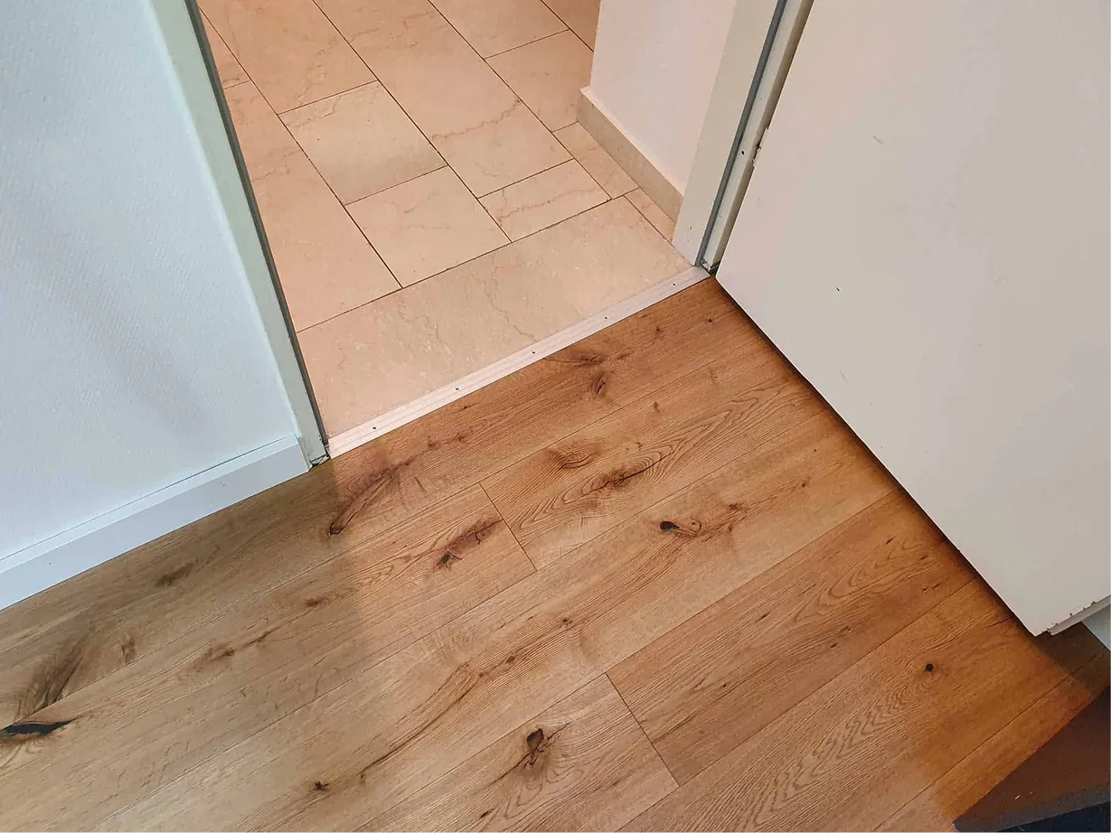 Türöffnung zwischen einem Boden mit hellbraunem Holzdesign und einem Fliesenboden in cremefarbenen Tönen.