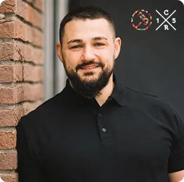 Ein Mann mit kurzem schwarzen Haar und Bart lehnt an einer Ziegelwand, trägt ein schwarzes Poloshirt und lächelt freundlich.