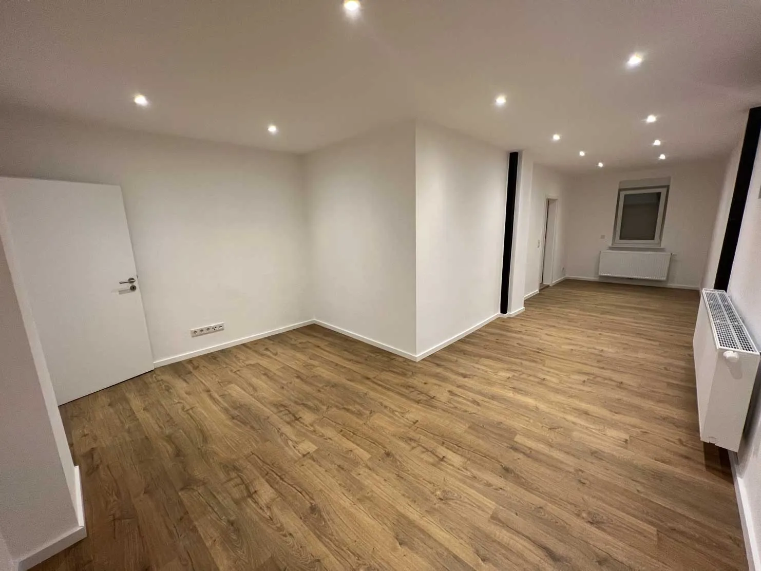 Leerer Raum mit weißen Wänden, Holzfußboden, mehreren Deckenleuchten, Fenster und Heizkörpern.