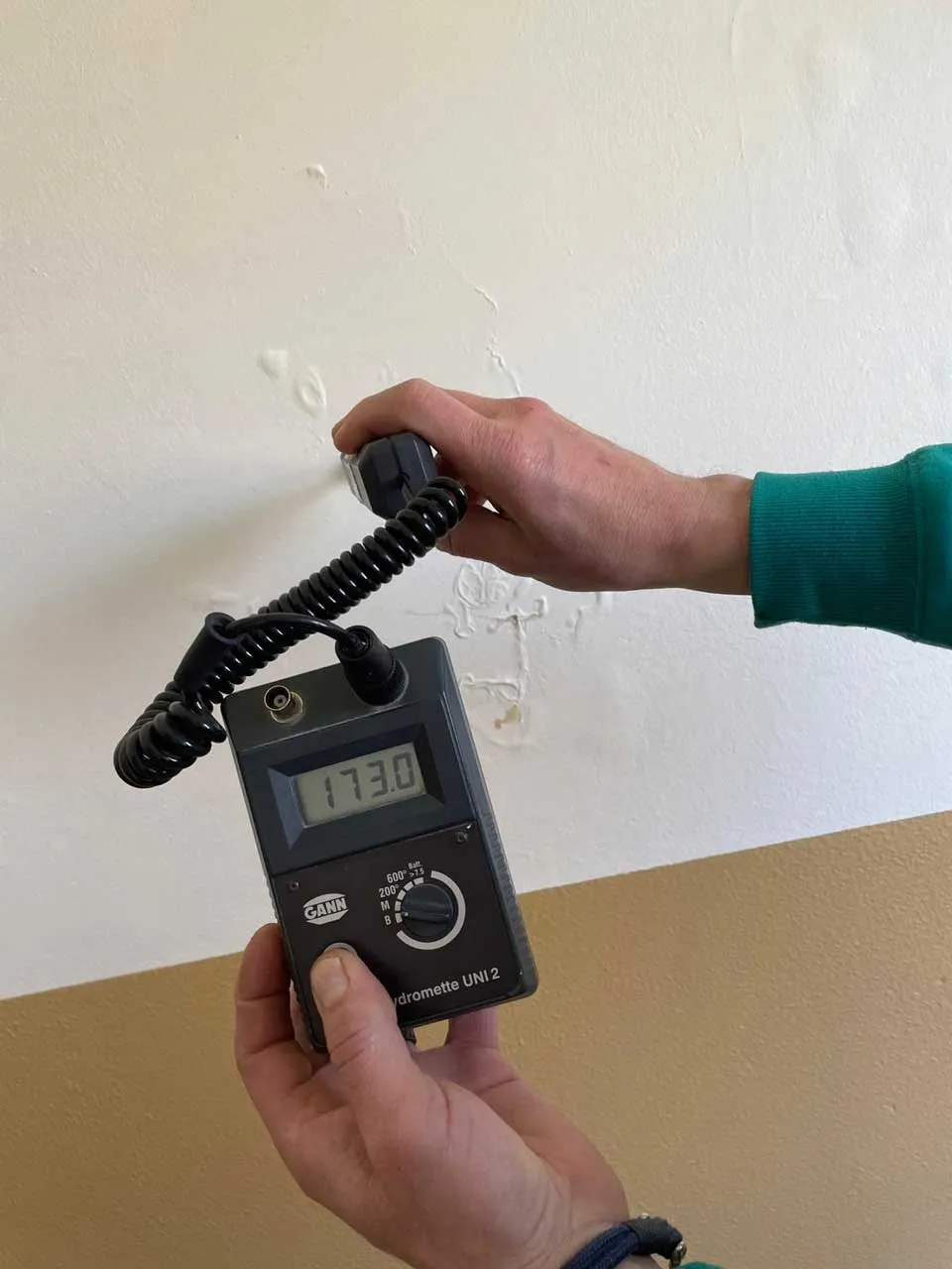 Person hält ein Hydrometer UNI 2 Messgerät gegen eine weiße Wand mit sichtbaren Feuchtigkeitsschäden.