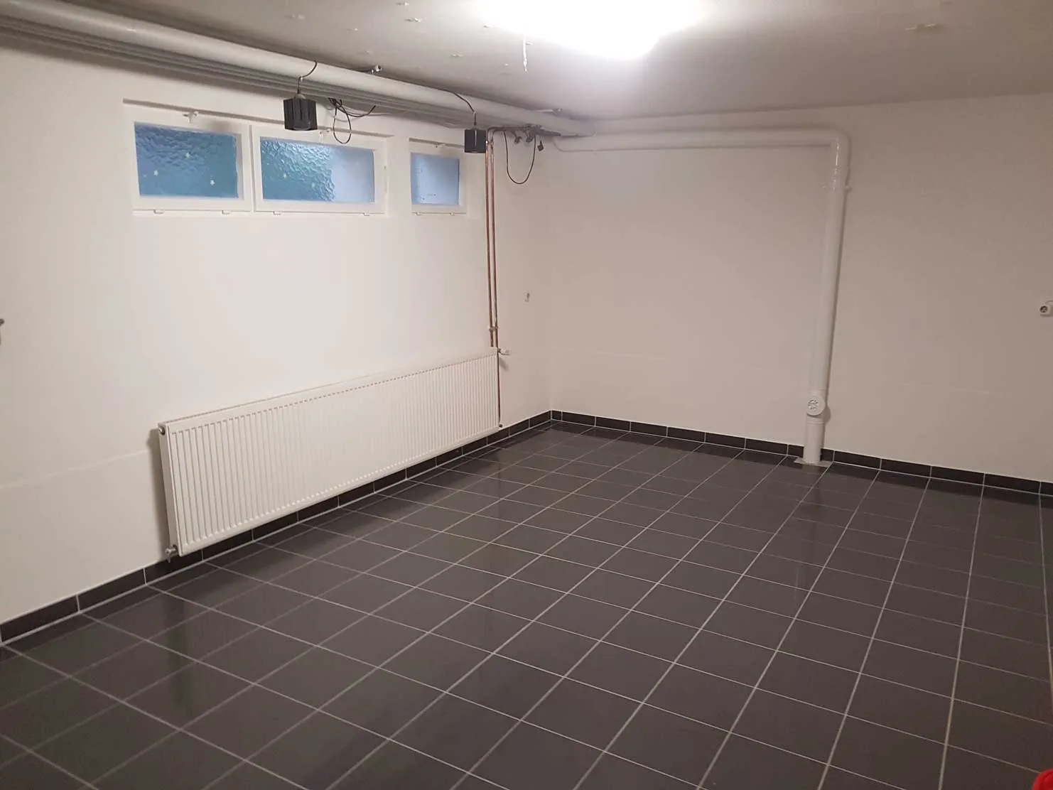 Leerer Raum mit grauen Fliesenboden, weißen Wänden, Heizkörper und drei kleinen Fenstern.