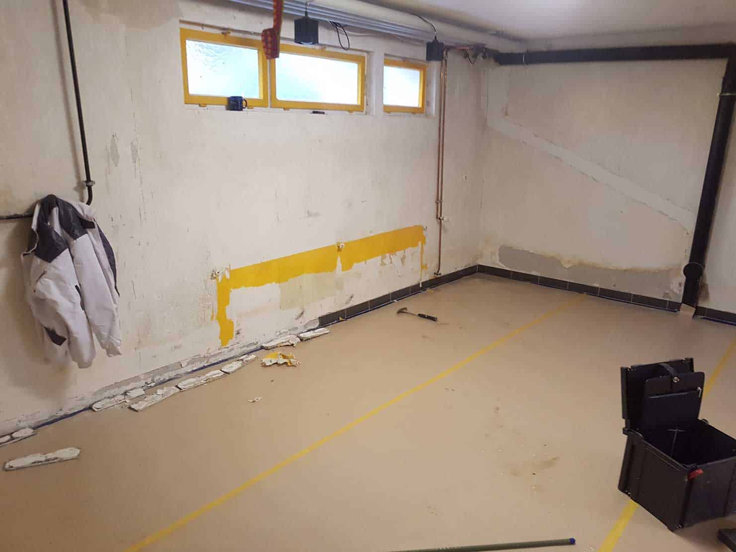 Leer stehender Kellerraum mit beschädigten Wänden, einer Jacke, Werkzeugkasten und teilweise abgeblätterter gelber Farbe an der Wand.