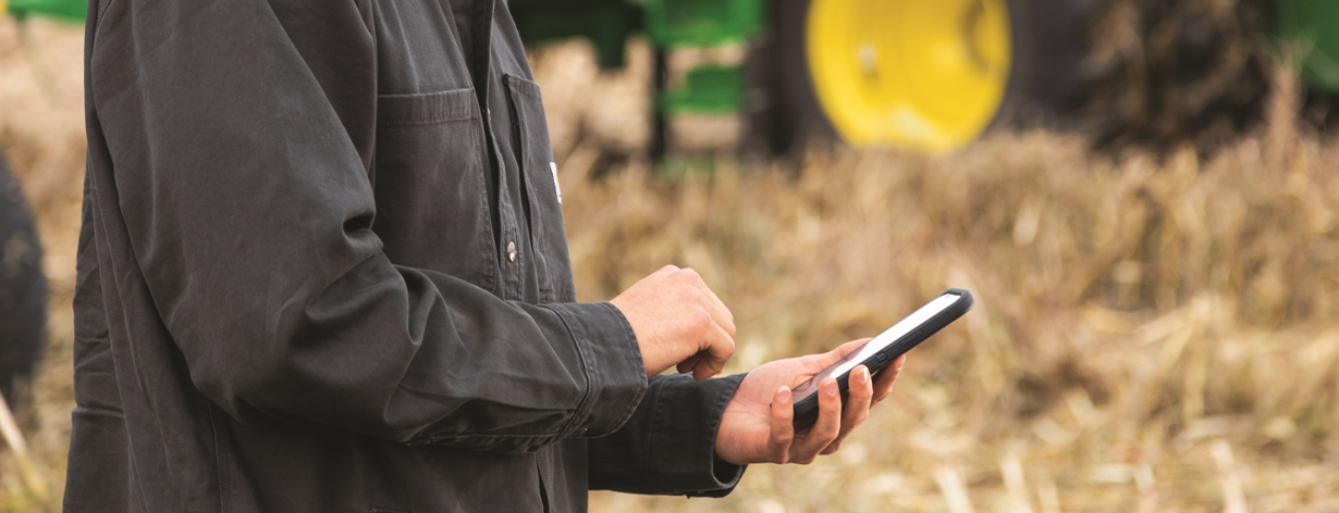 Person wearing a dark jacket using a smartphone outdoors with blurred farm equipment in the background.