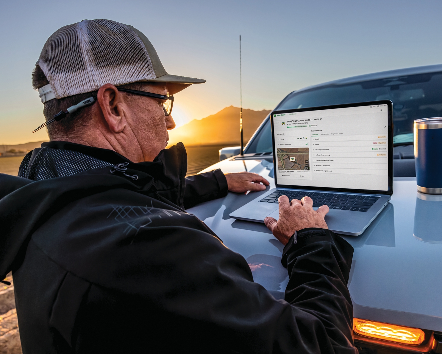 Man wearing a cap and jacket using a laptop on the hood of a vehicle at sunset.
