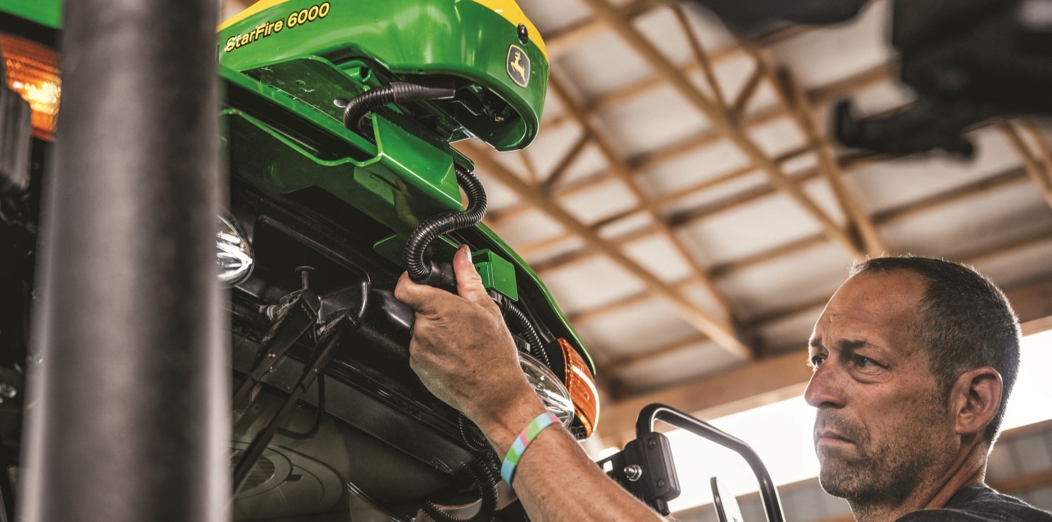 Man inspecting wiring on a green John Deere StarFire 6000 device inside a barn.
