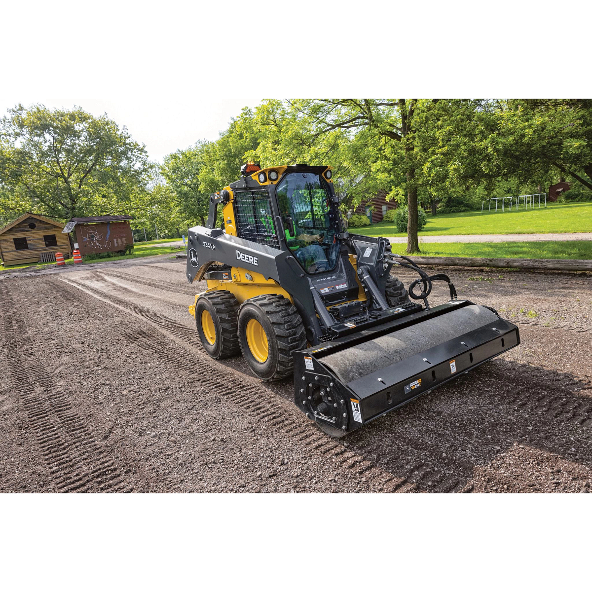 334 P-Tier skid steer loader equipped with a large cylindrical brush attachment cleans and smooths a dirt surface in a park-like setting.