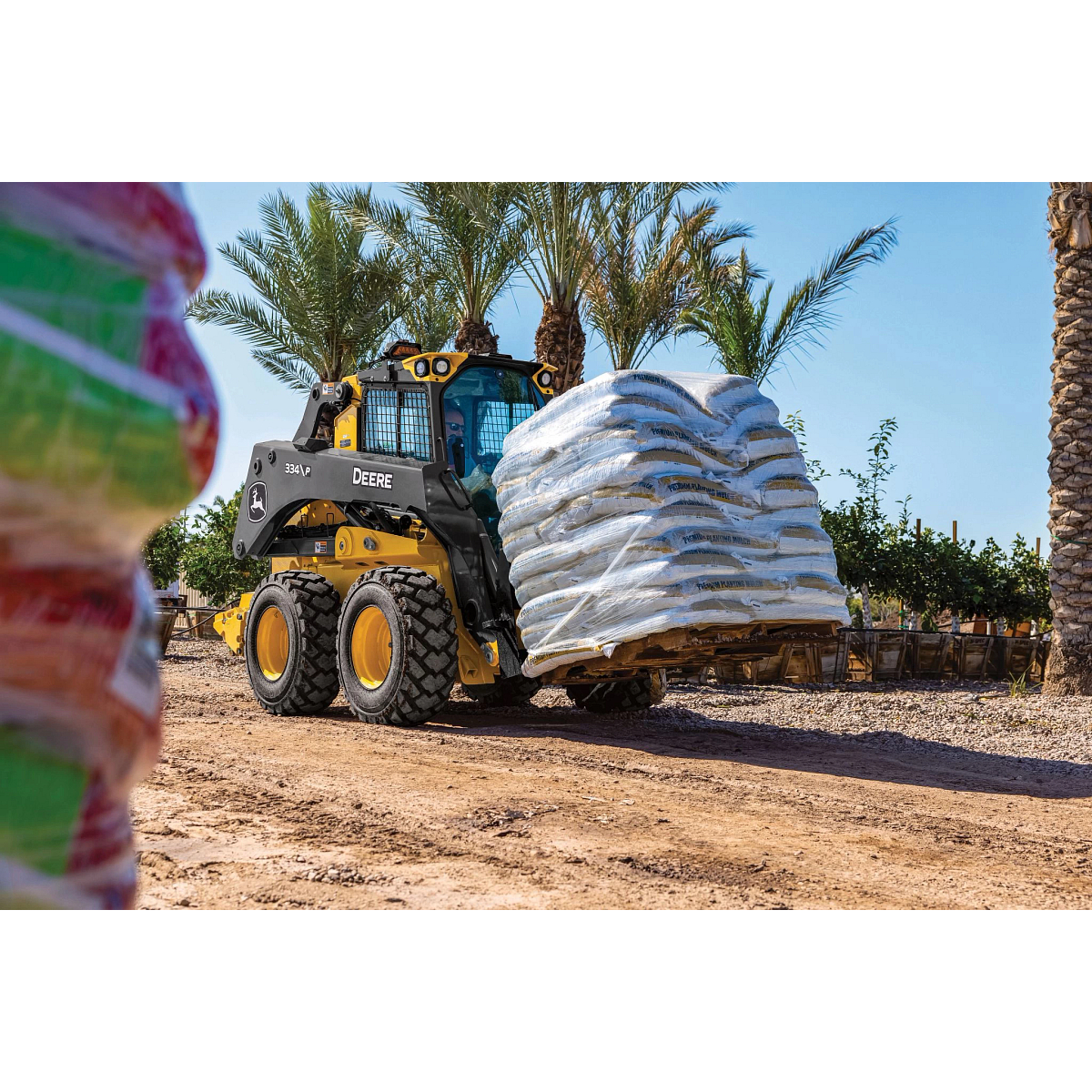 334 P-Tier skid steer loader transports a large pallet stacked with white bagged materials along a dirt path.