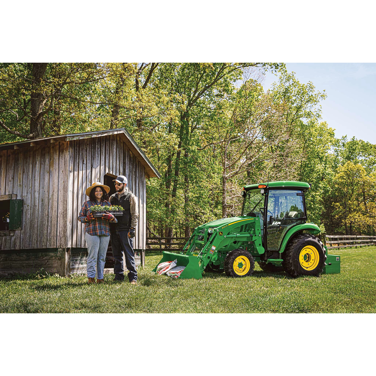 Two farmers standing in front of a wood building in front of a fenced-off field and woods, with a green 3046R compact utility tractor parked to the right of them.