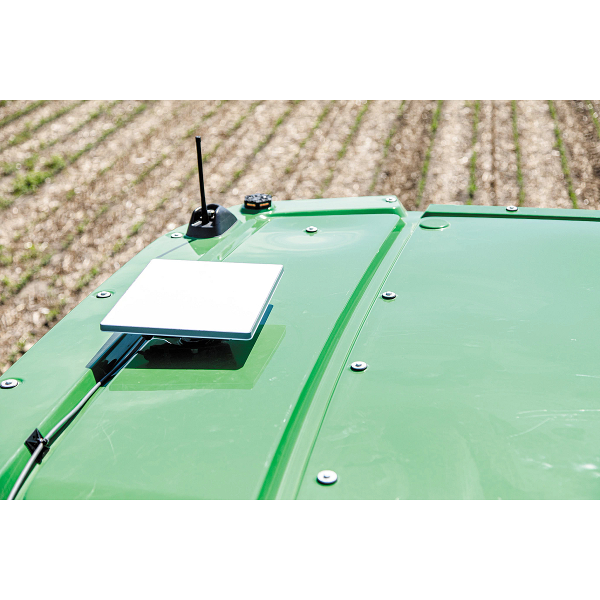 JDLink Boost Mini Terminal mounted on the roof of a John Deere machine beside an antenna, with cultivated crop rows visible in the background.