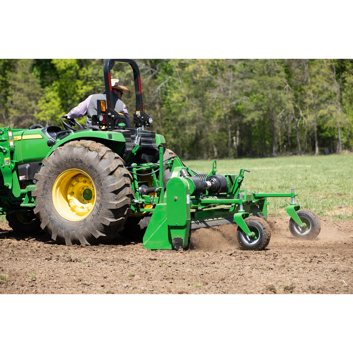 Tractor tilling a field, with soil turning behind the implement and trees in the background.