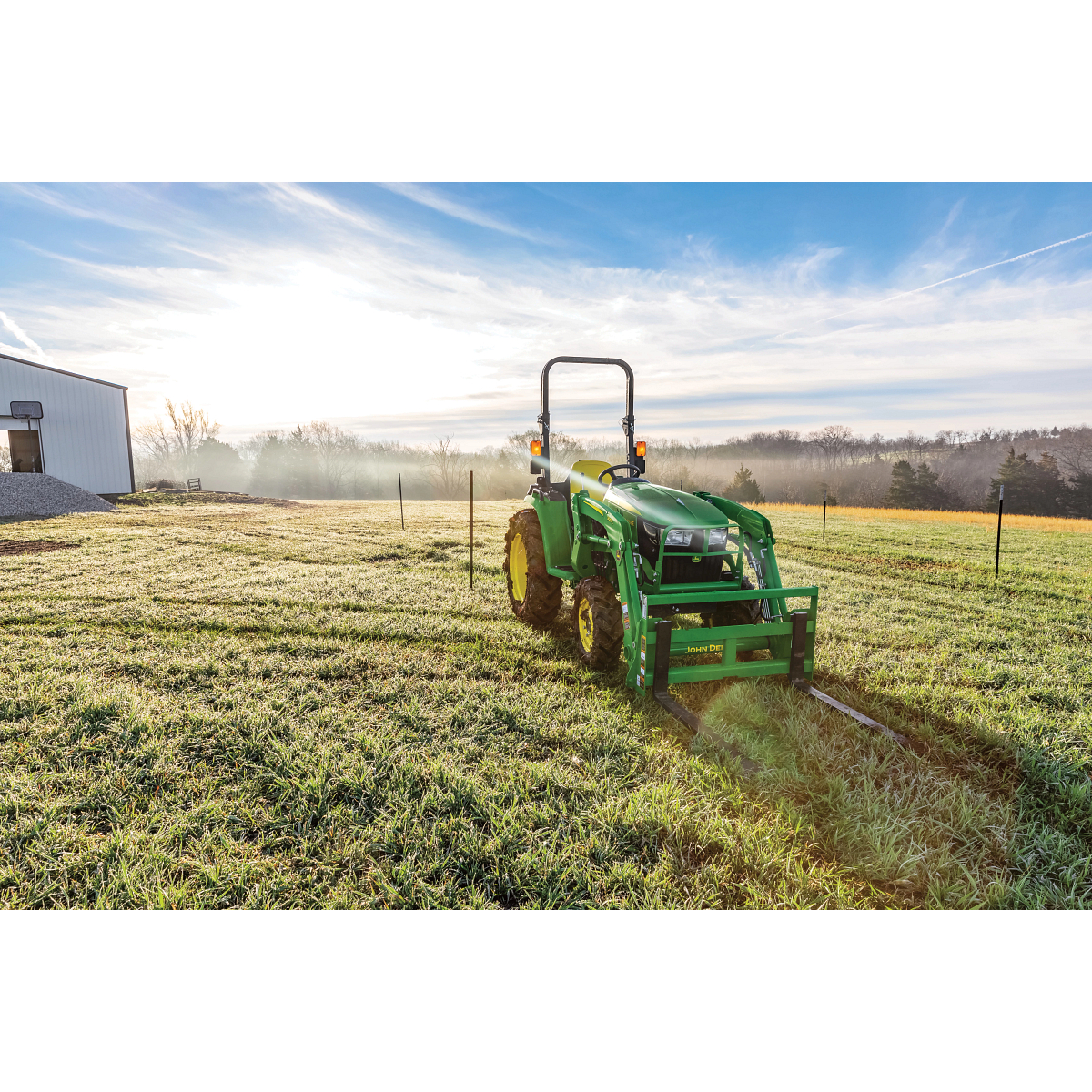 Green tractor parked in a frosty grass field as a building sits to the left under a bright early morning sky, with long shadows stretching across the ground and rolling landscapes in the background