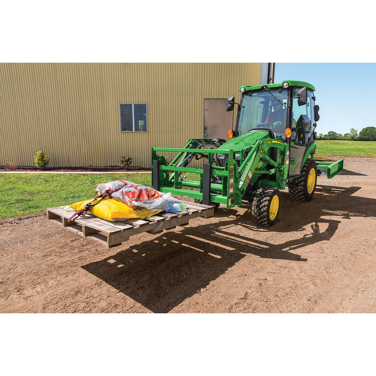 Green tractor with pallet forks lifts a loaded wooden pallet beside a metal building on a gravel surface, with open farmland visible in the background under clear sky