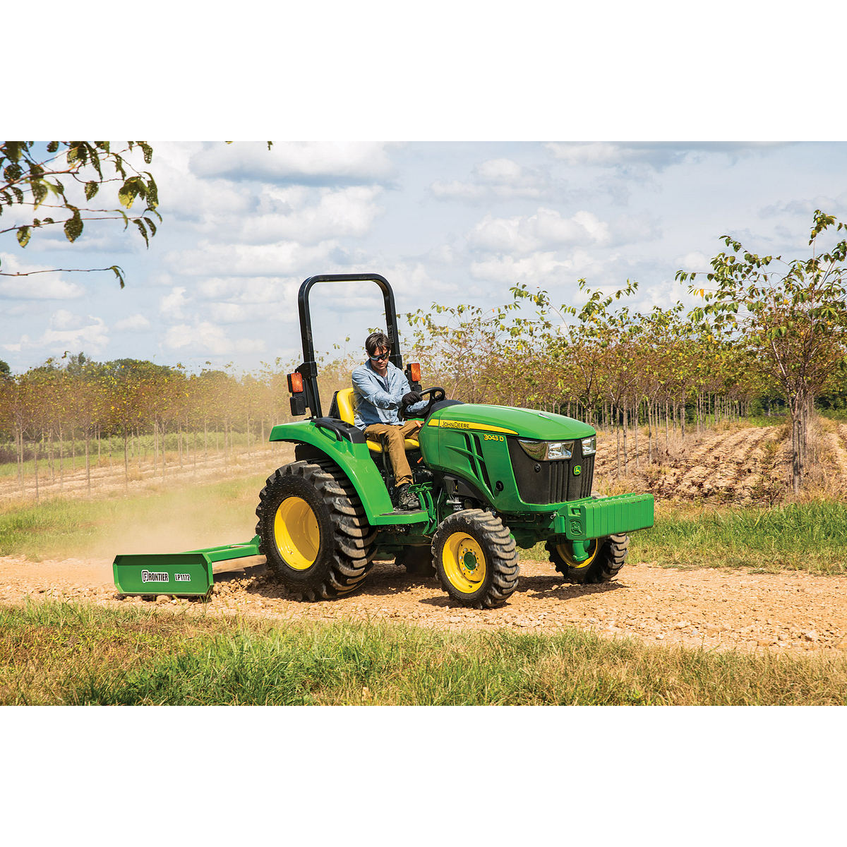 A person drives a green tractor pulling a land plane attachment along a dirt path beside rows of trees under a partly cloudy sky