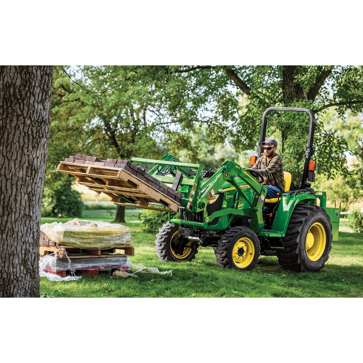 Front left view of a 3032E compact tractor lifting a pallet of brick with an AP11F pallet fork on a shady, green lawn