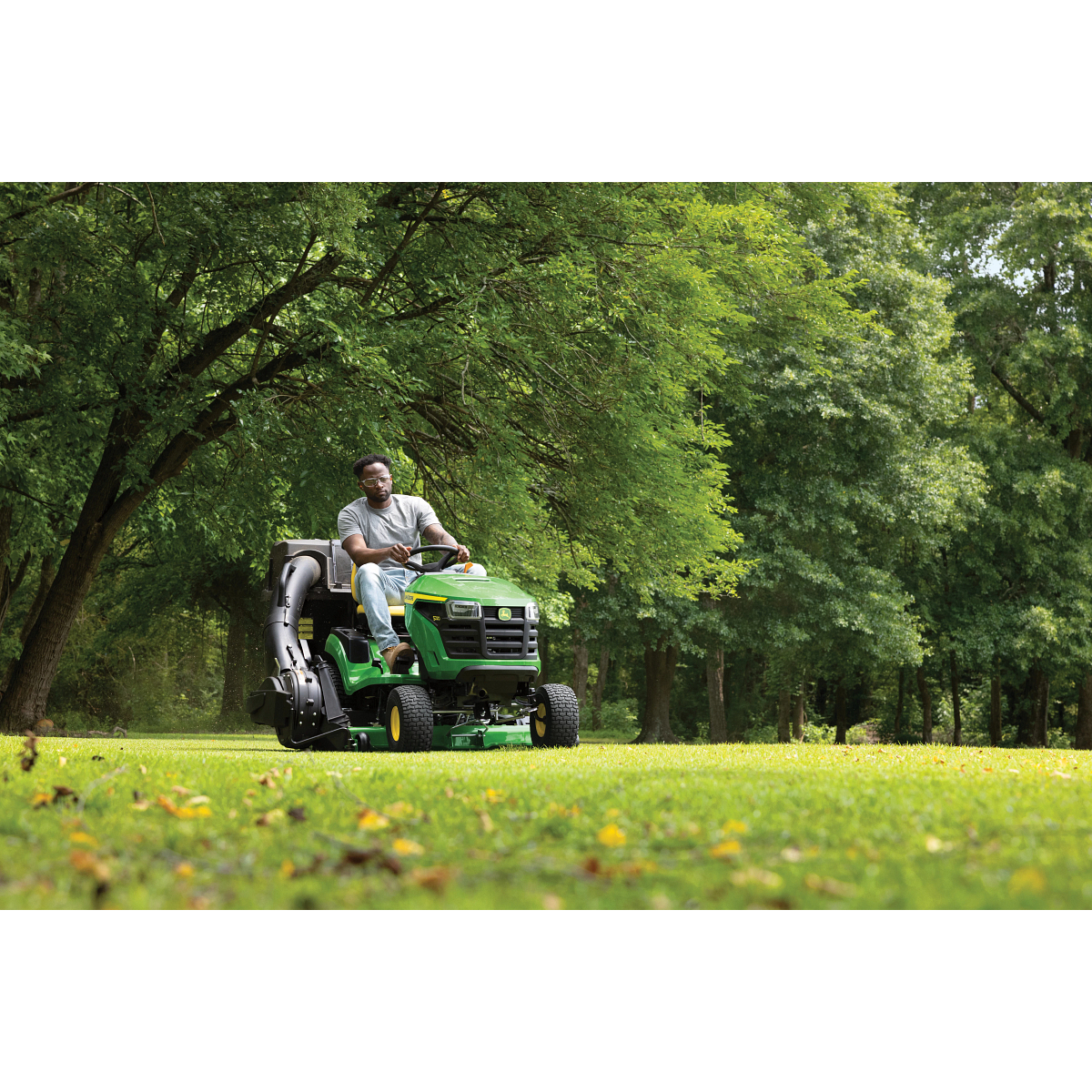 Front right view of an operator using an S220 Lawn Tractor with attached material collection system on a wooded lot