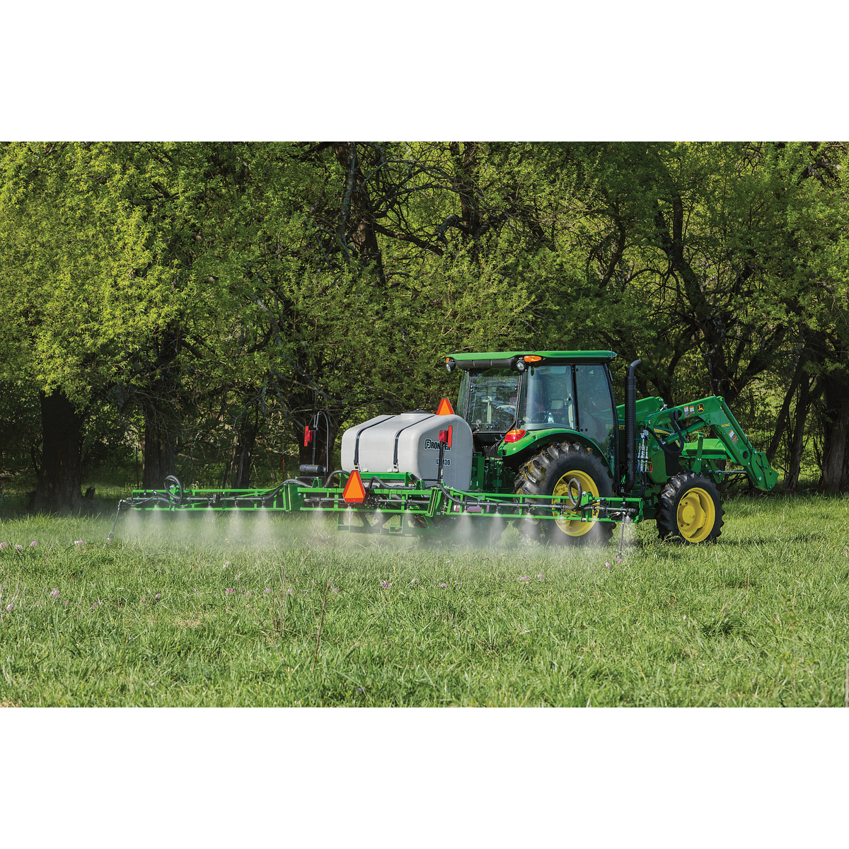 A tractor with a boom sprayer drives across a grassy field, spraying a fine mist over the crop, with leafy trees forming a dense green background.