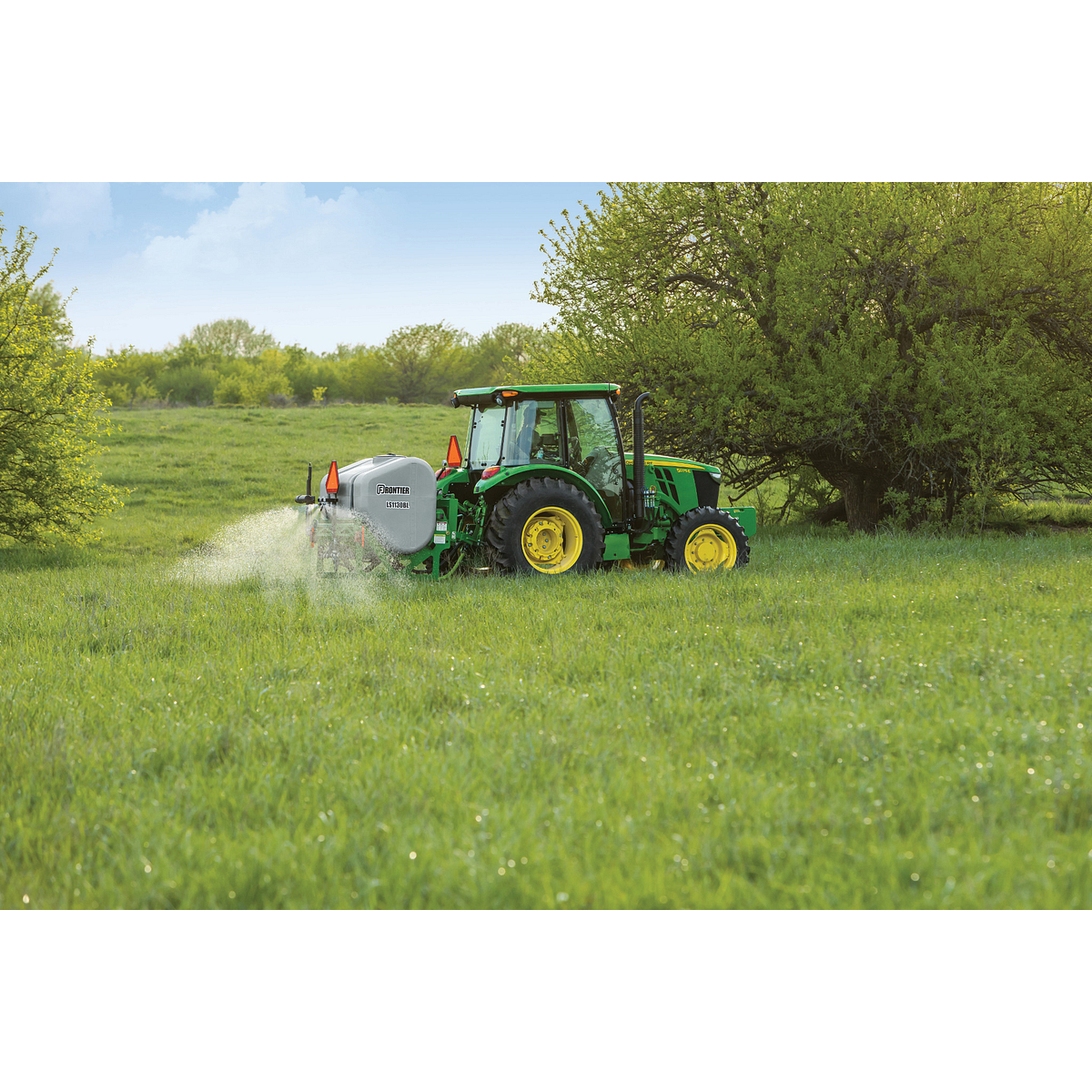 A tractor pulls a sprayer across a lush, grassy field, releasing a fine mist near a tree line on a sunny day.