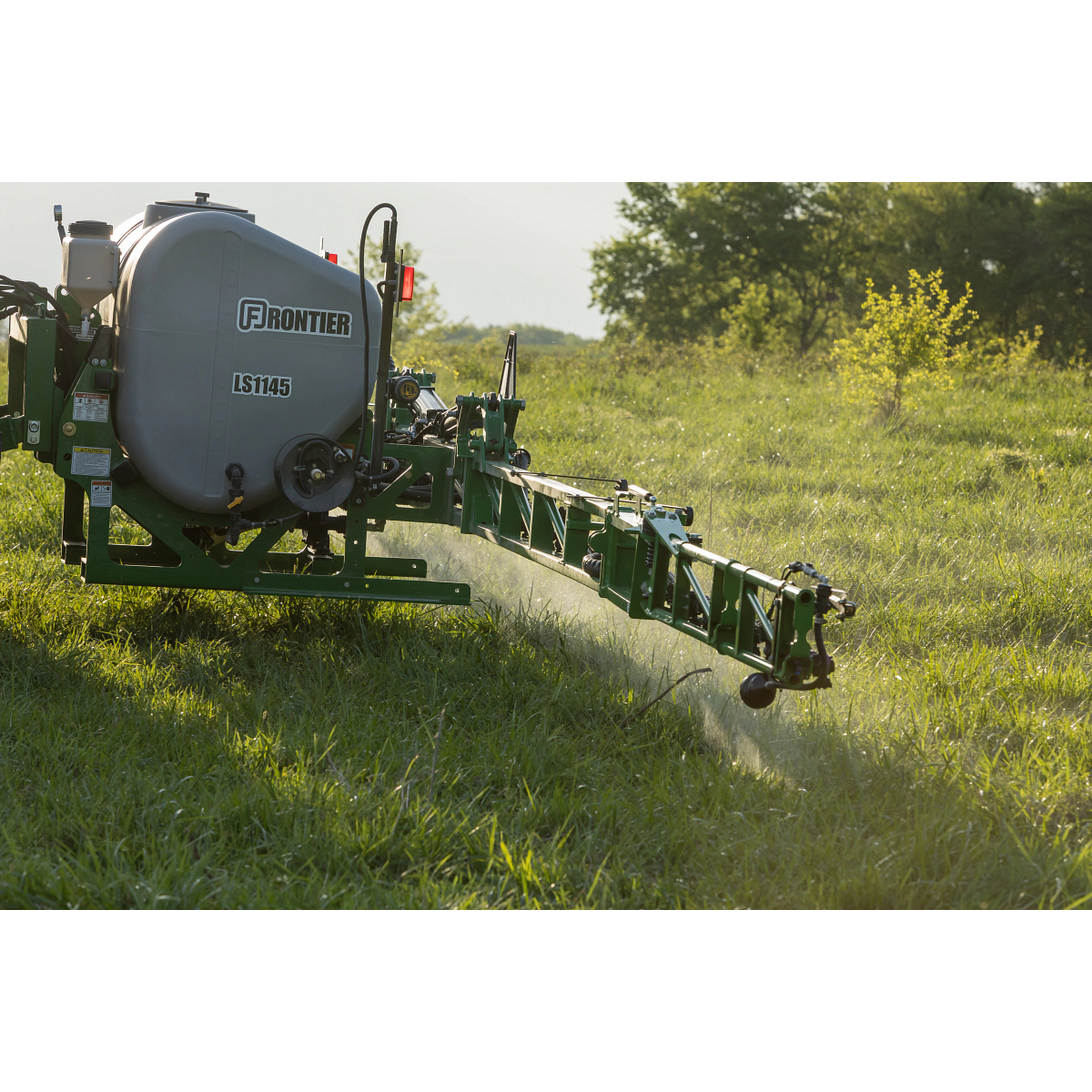 Rear-mounted agricultural sprayer labeled Frontier LS1145 with boom extended, spraying liquid over tall grass in a sunny field with trees in the background.